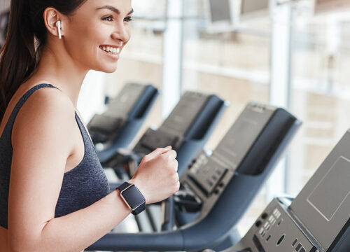 Side view of young and cheerful woman in sportswear is running on a treadmill at gym and listening music.