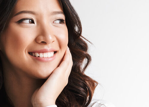 Image of smiling woman with long dark hair looking aside at copyspace and touching cheek isolated over white background in studio